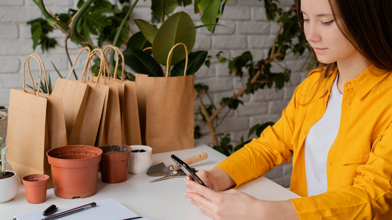Женщина в желтой рубашке смотрит на свой телефон за столом с растениями и бумажными пакетами.